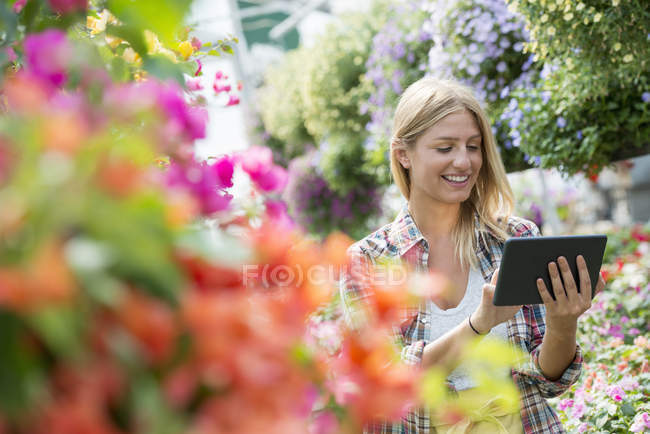 Young woman examining flowers with digital tablet in plant nursery. — Stock Photo