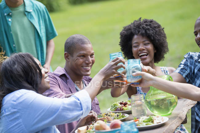 Giovani amici tifo con bevande al tavolo da picnic nel giardino di campagna . — Foto stock