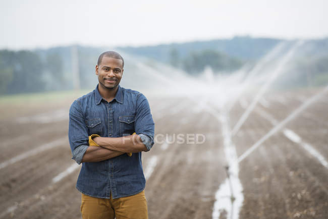 Giovane agricoltore di sesso maschile in abiti da lavoro su campo biologico con irrigazione irrigatori ad acqua . — Foto stock