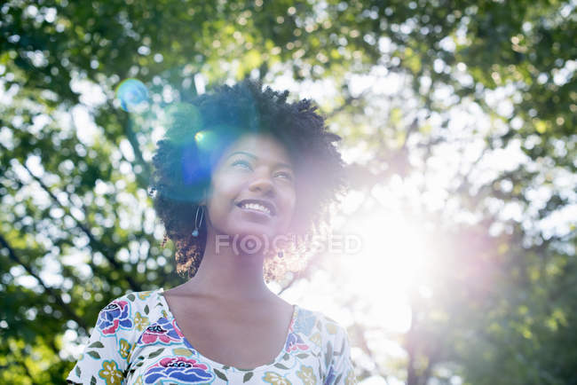 Giovane donna in abito fiorito sorridente e guardando in alto nella foresta soleggiata . — Foto stock