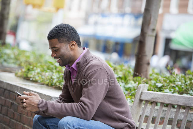 Giovane seduto sulla panchina e controllare il telefono in città
. — Foto stock