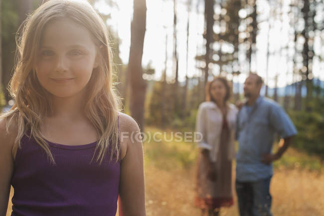 Chica de edad elemental con el pelo rubio de pie en el bosque con adultos en el fondo . - foto de stock