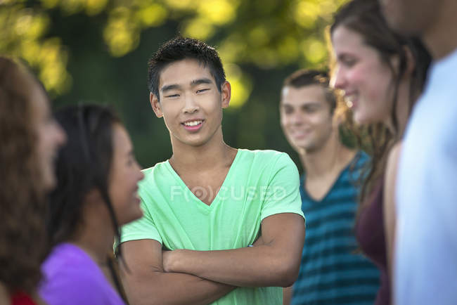 Teenage boy standing with arms crossed in group of young laughing friends outdoors. — Stock Photo
