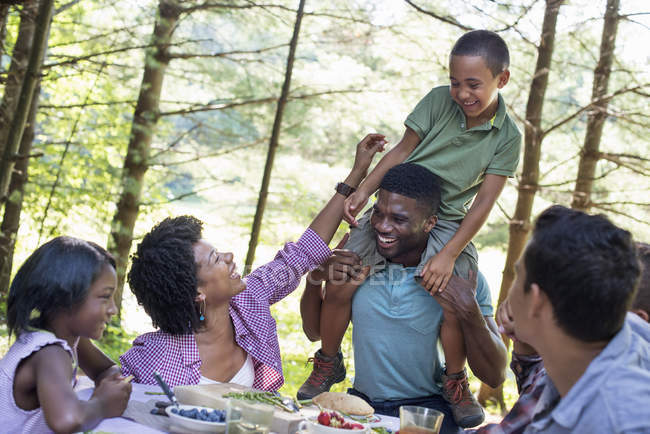 Elementary age boy sitting on shoulders of young man by picnic table with family in forest. — Stock Photo
