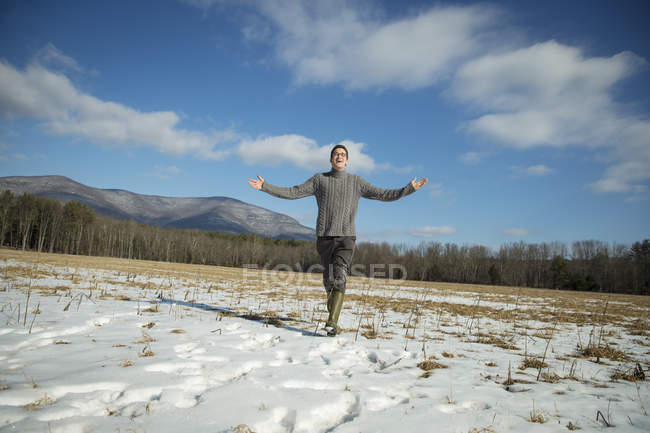 Man in knit jumper and muck boots standing with arms outstretched in snowy rural landscape. — Stock Photo