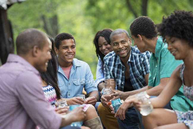 Joyeux jeunes amis prenant des boissons, parlant et riant dans le jardin de campagne . — Photo de stock