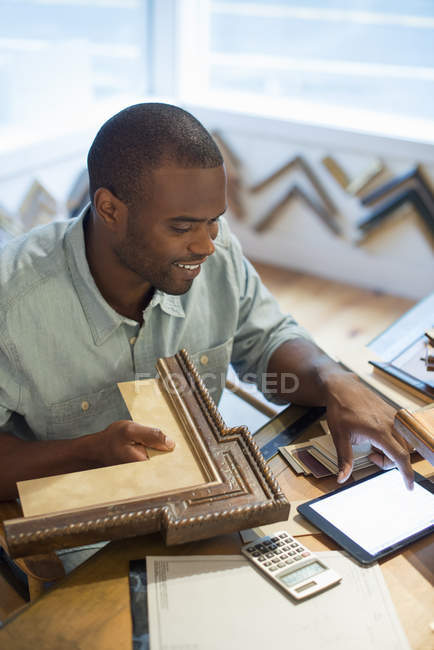 Young man holding picture frame and using digital tablet at workbench in picture framing studio. — Stock Photo