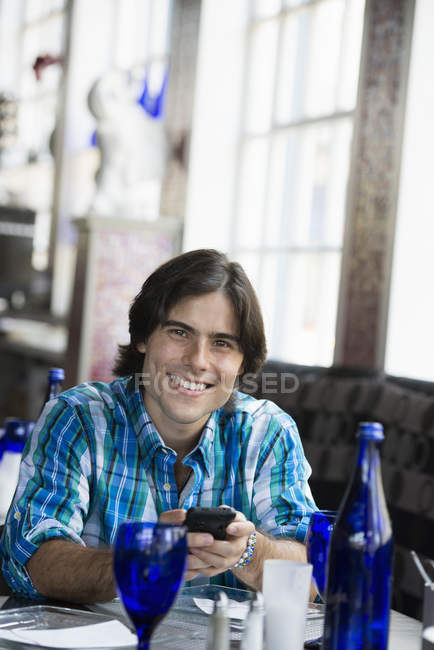 Hombre joven sentado en la mesa en la cafetería y el uso de teléfono inteligente . - foto de stock