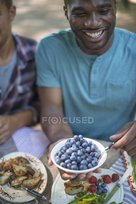 Young man holding bowl of fresh blueberries at picnic table with family in woods. — Stock Photo