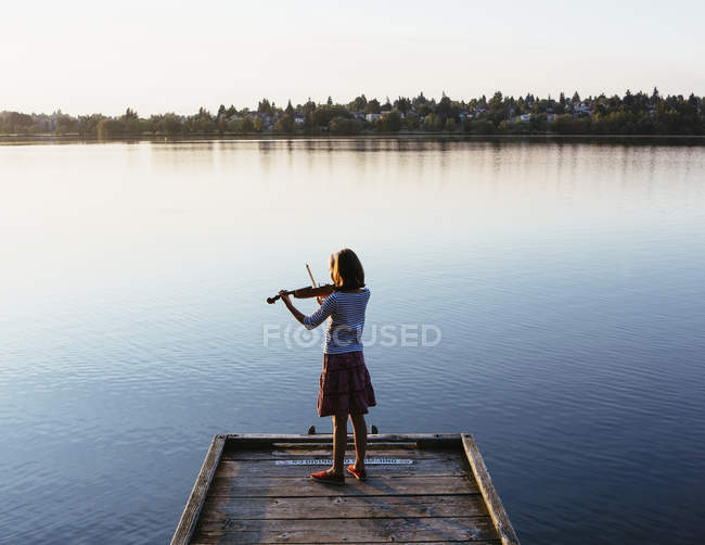 Pre-adolescent female violinist playing violin at dawn on wooden pier at lake. — Stock Photo