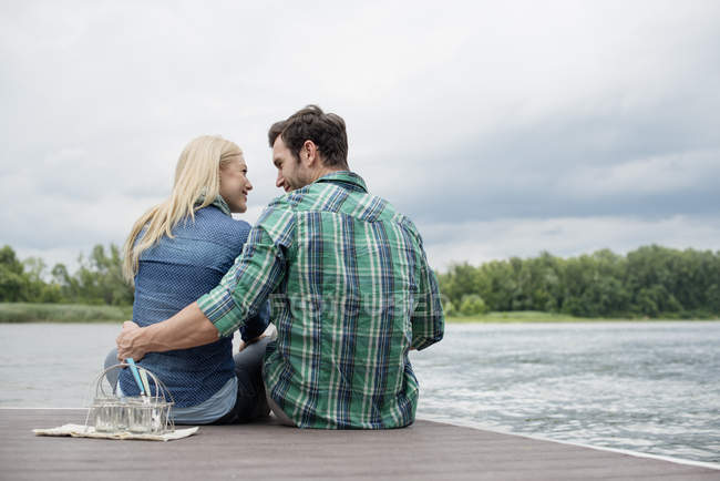 Man and woman sitting head to head and embracing on jetty with glasses by lake. — Stock Photo