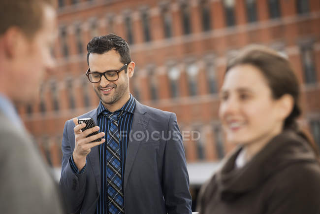 Mid adult businessman using smartphone with people talking in foreground. — Stock Photo