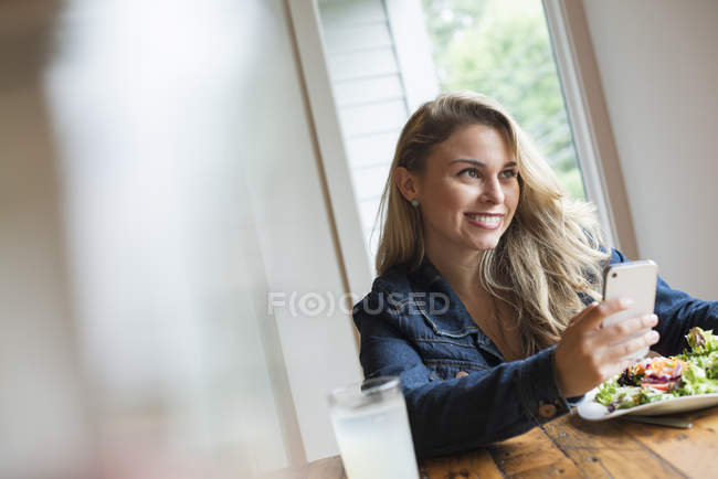 Young woman smiling while using smartphone at cafe table with drink and meal. — Stock Photo