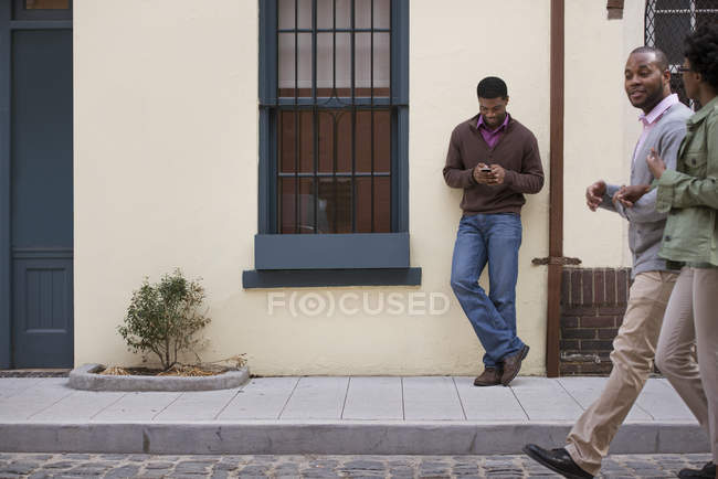 Casal andando na calçada com o homem encostado à parede e verificando o telefone . — Fotografia de Stock