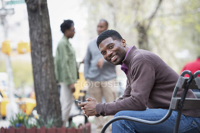 Giovane seduto sulla panchina della città con le persone che parlano in background . — Foto stock