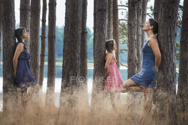 Tres mujeres jóvenes apoyadas en árboles en el bosque con lago . - foto de stock