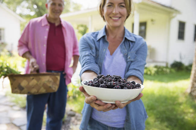 Mature couple posing with fresh blackberries and vegetables at farmhouse green garden. — Stock Photo
