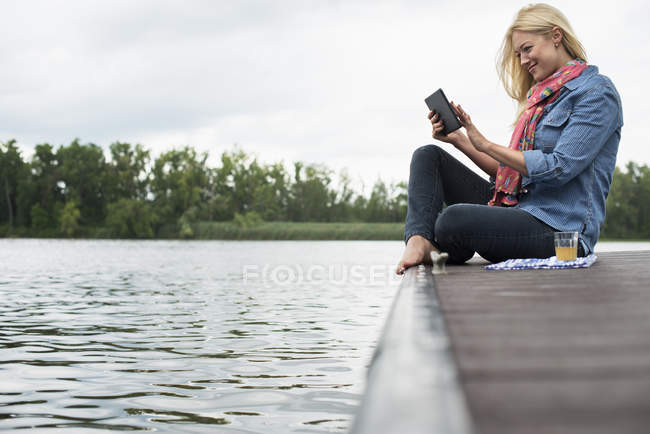 Side view of woman sitting on jetty by lake and using digital tablet. — Stock Photo