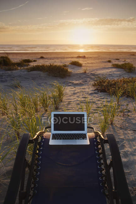 Ordenador portátil en silla de cubierta con vistas a la puesta del sol en la playa - foto de stock