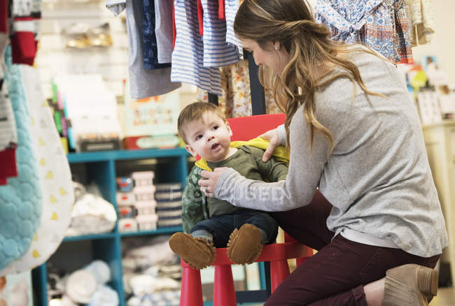 Кавказская мать и маленький сын сидят на стуле во время покупок в магазине одежды — стоковое фото
