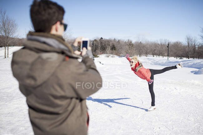 Joven hombre tomando foto de la mujer patinaje sobre hielo en el lago congelado en invierno - foto de stock