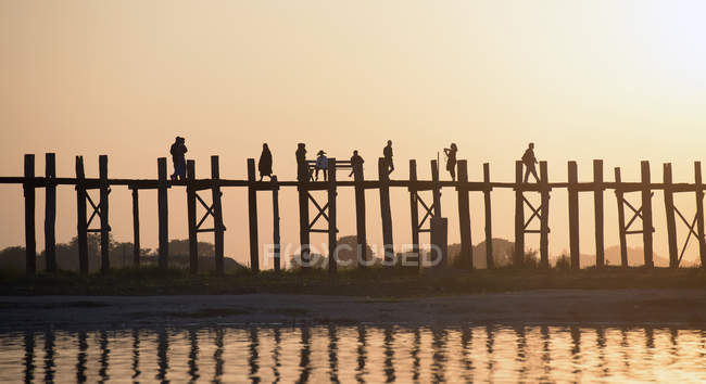 Personas caminando por una pasarela de madera elevada al atardecer en Myanmar - foto de stock