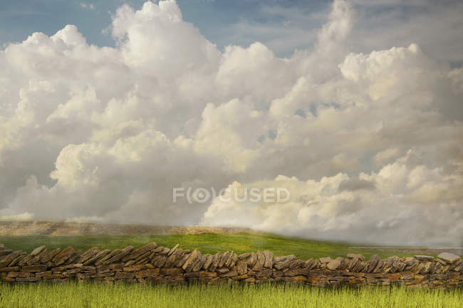 Muralla de piedra y campo de hierba bajo las nubes en el paisaje rural - foto de stock