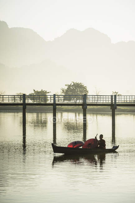 Montanhas e reflexão ponte em lago imóvel com barco com monge budista jovem e guarda-chuvas, Hpa-an, Kayin, Mianmar — Fotografia de Stock