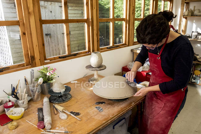 Frau in roter Schürze steht in Keramikwerkstatt und arbeitet an Tonschale. — Stockfoto