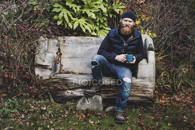 Uomo barbuto che indossa berretto nero seduto su una panchina di legno in giardino, tenendo la tazza blu, guardando in macchina fotografica
. — Foto stock