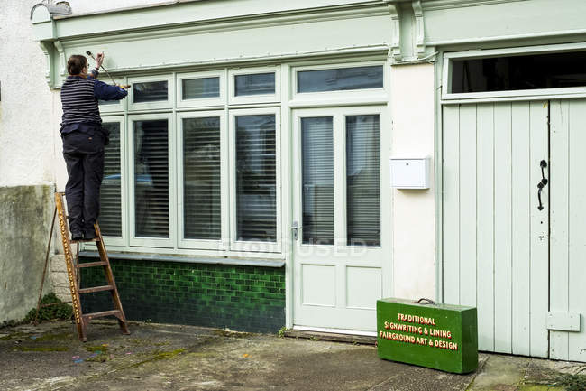 Woman standing on ladder and using paintbrush and maulstick while working on sign-writing architrave above shop window. — Stock Photo