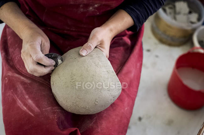 Midsection of ceramic artist in red apron sitting in workshop, working on clay vase. — Stock Photo