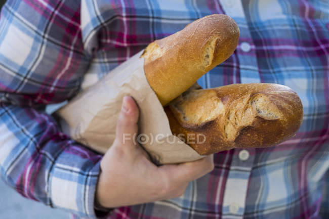 Close up of person holding two freshly baked French baguettes in brown paper bag. — Stock Photo