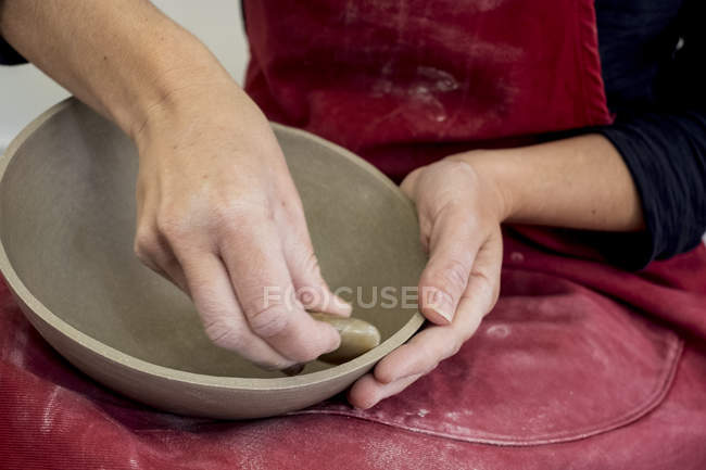 Sección media del artista de cerámica en delantal rojo trabajando en un tazón de barro pequeño . - foto de stock