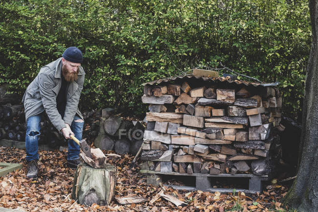 Homme barbu en bonnet noir et parka debout dans le jardin en automne, en utilisant la hache pour couper un morceau de bois sur le bloc de coupe . — Photo de stock