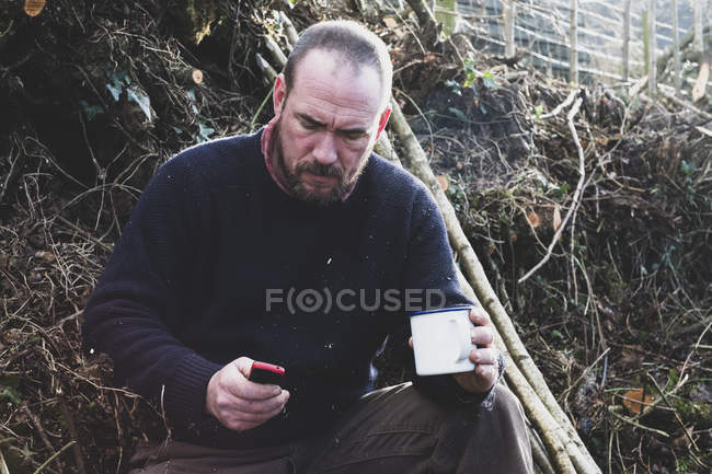 Homem barbudo sentado no chão ao lado de um monte de estacas de madeira, segurando caneca, verificando o telefone celular . — Fotografia de Stock