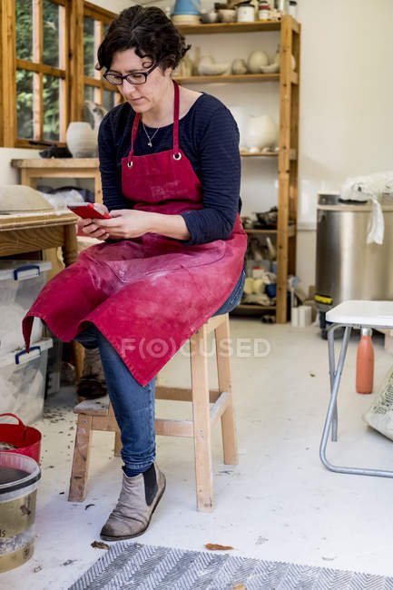 Жінка в червоному фартусі сидить на табуреті в керамічній майстерні і перевіряє мобільний телефон . — стокове фото