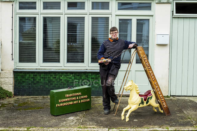 Female signwriter holding paintbrush, palette and mahlstick beside ladder and small carousel horse. — Stock Photo