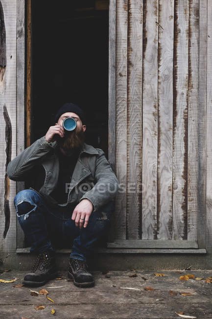 Homem barbudo sentado na porta da oficina de madeira, bebendo chá de caneca azul . — Fotografia de Stock