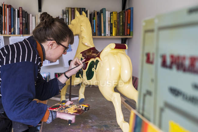 Woman standing in workshop and painting traditional wooden carousel horse from merry-go-round. — Stock Photo