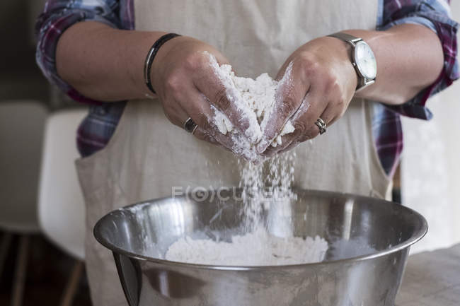 Close up of person wearing apron standing in kitchen, mixing ingredients for crumble in metal bowl. — Stock Photo