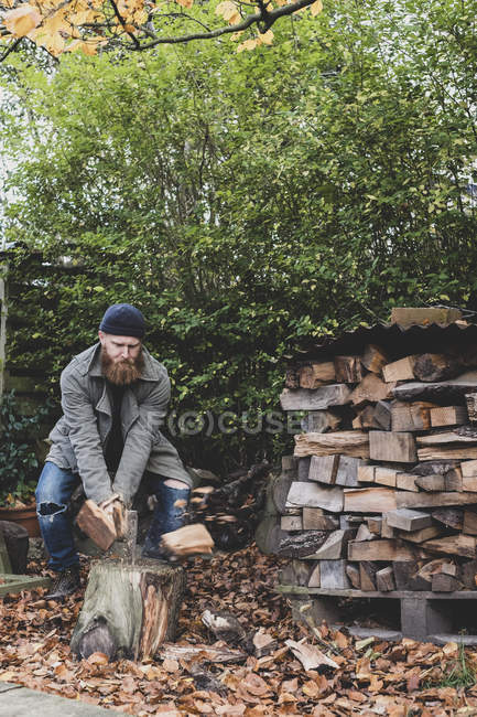 Bearded man in black beanie and parka standing in garden in autumn, using axe to chopping piece of wood on chopping block. — Stock Photo