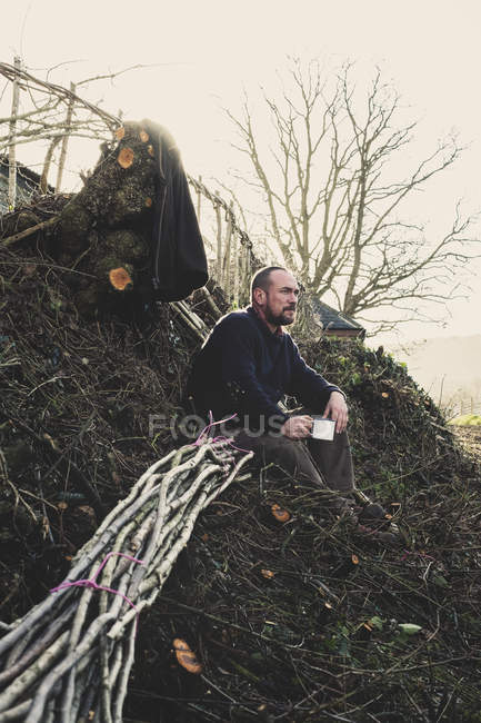 Bärtiger Mann sitzt auf dem Boden neben einem Haufen hölzerner Bittsteller, die im traditionellen Heckenbau verwendet werden. — Stockfoto