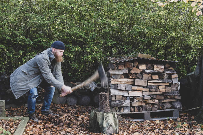 Homme barbu en bonnet noir et parka debout dans le jardin en automne, en utilisant la hache pour couper un morceau de bois sur le bloc de coupe . — Photo de stock