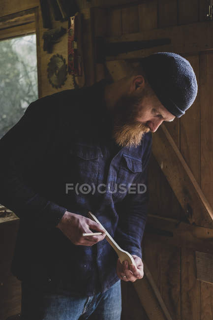 Homem barbudo vestindo gorro preto em pé na oficina, examinando pedaço de madeira . — Fotografia de Stock