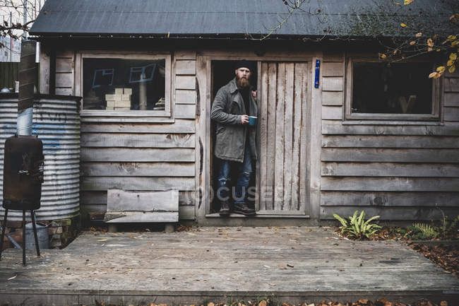 Bärtiger Mann steht vor Tür der Holzwerkstatt, hält blauen Becher in der Hand und blickt in die Kamera. — Stockfoto