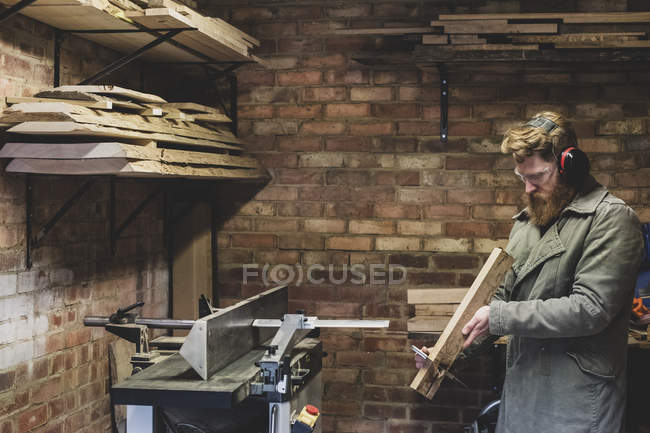 Uomo barbuto in piedi in officina, indossando protezioni per le orecchie, lavorando su un pezzo di legno . — Foto stock