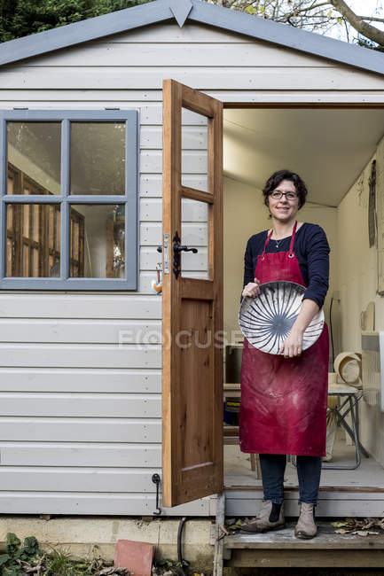 Woman in red apron standing on steps outside workshop and holding ceramic bowl with black lines pattern. — Stock Photo