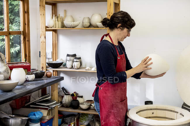 Mulher de avental vermelho em pé na oficina ao lado do forno e segurando vaso de cerâmica
. — Fotografia de Stock