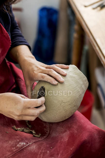 Manos de artista de cerámica en delantal rojo sentado en el taller, trabajando en jarrón de arcilla . - foto de stock